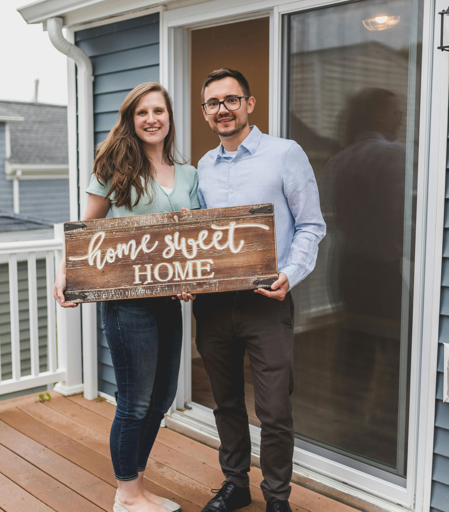 Smiling couple holding 'Home Sweet Home' sign in front of their new house.